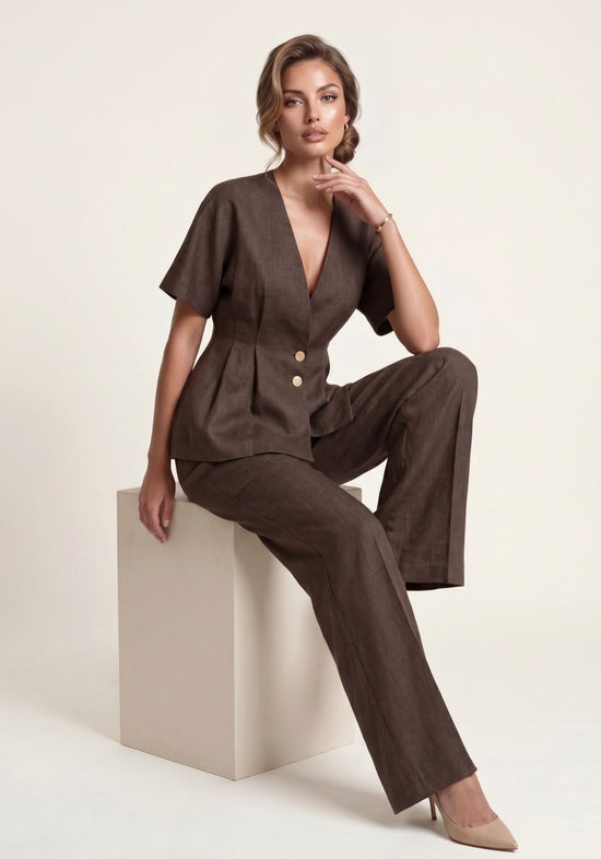 Brown women’s linen set, worn seated with blazer and wide trousers on a light pedestal.
