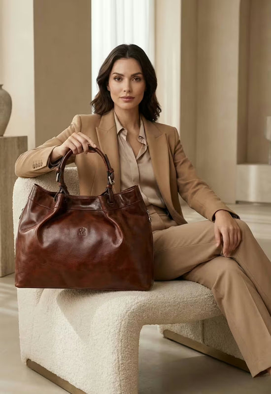 Brown leather handbag worn by a model while she sits on a light chair.