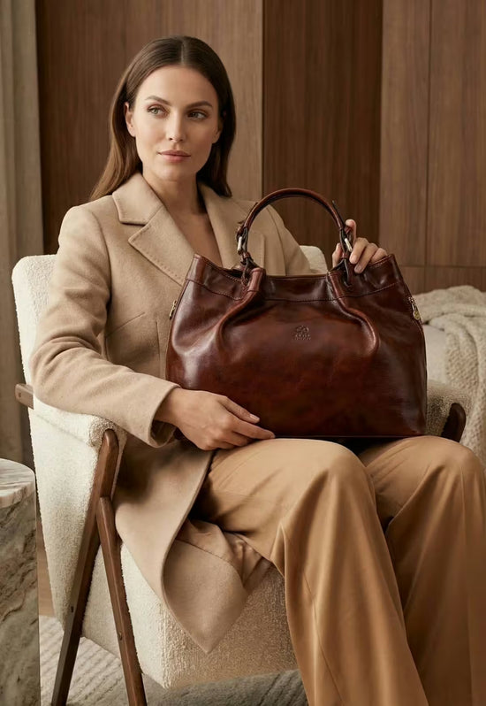 Brown leather handbag seen from the front, held and worn by the model in an armchair.