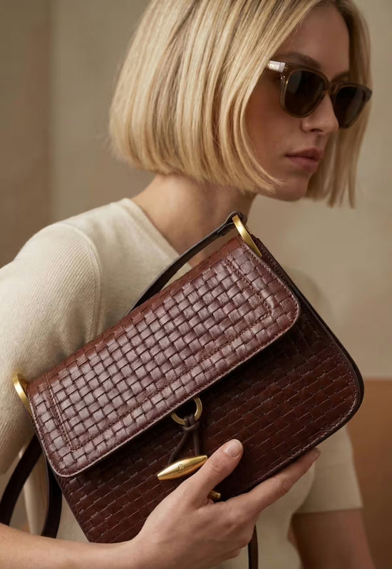 Women’s leather handbag in dark brown with flap, worn on the arm and shown close up.