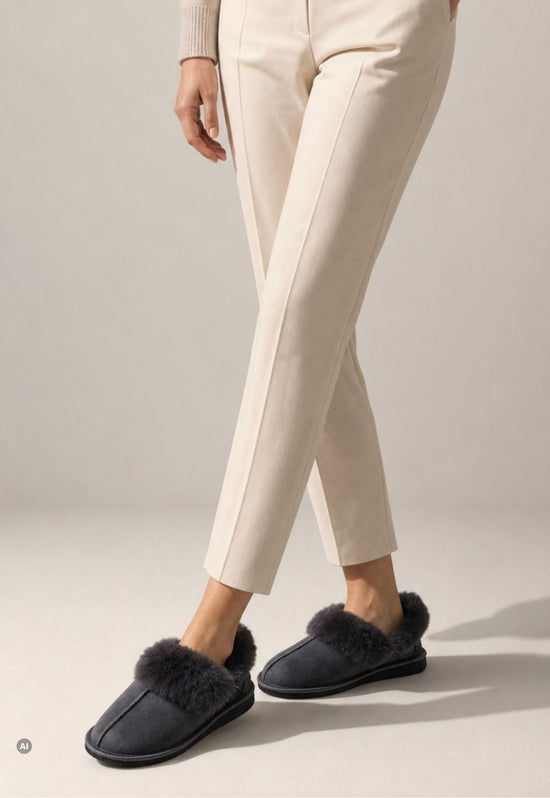 Women’s sheepskin slippers in anthracite, worn with light trousers and a visible fur collar.