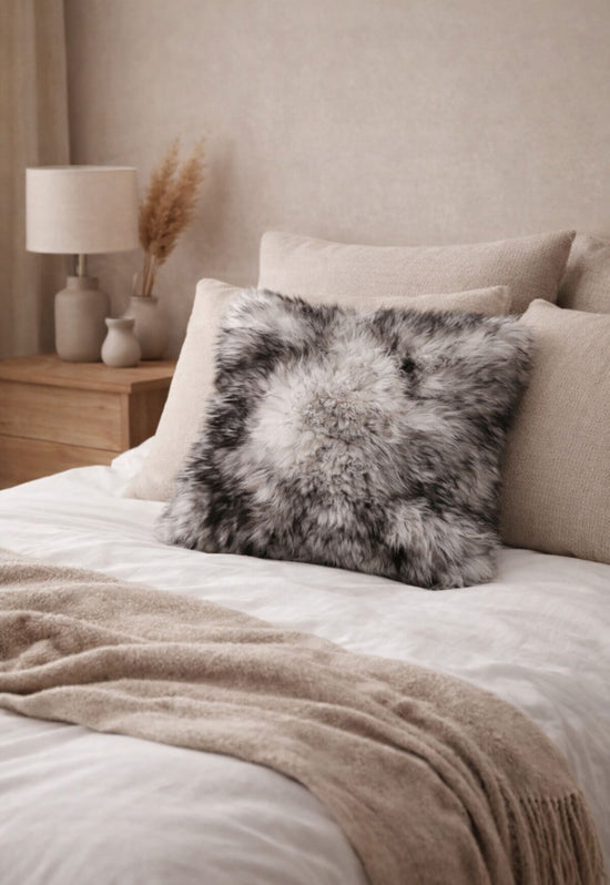 Grey sheepskin cushion on a bed with pale pillows and a throw.