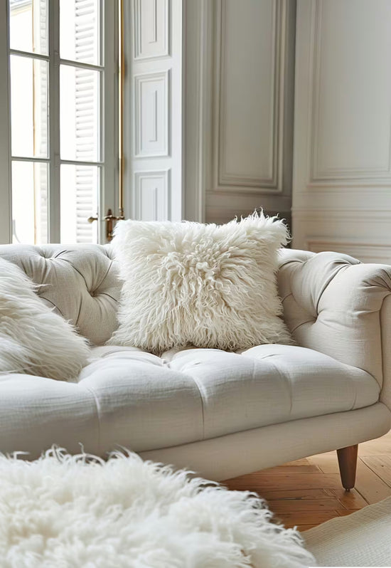 White sheepskin cushion as a detail on a tufted sofa in front of tall windows and light wood flooring.