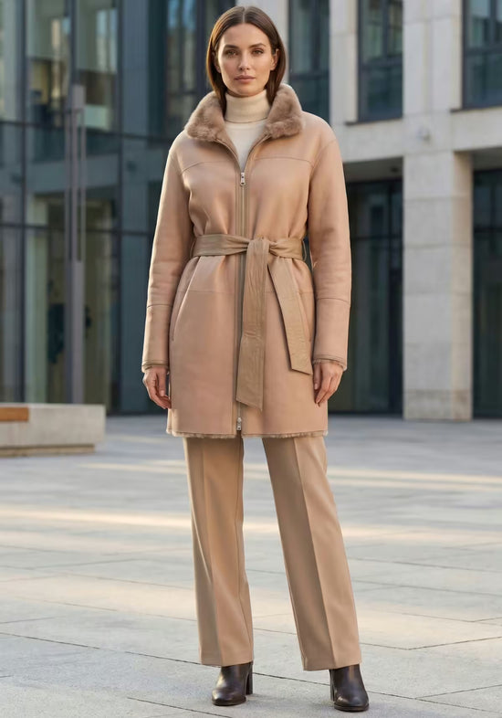 Beige women's sheepskin coat in a worn styling shot, open over trousers in an urban setting.