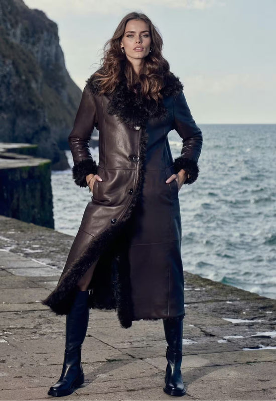 Women's sheepskin coat in dark brown as a worn image by the coast, worn open with a belt and fur collar.