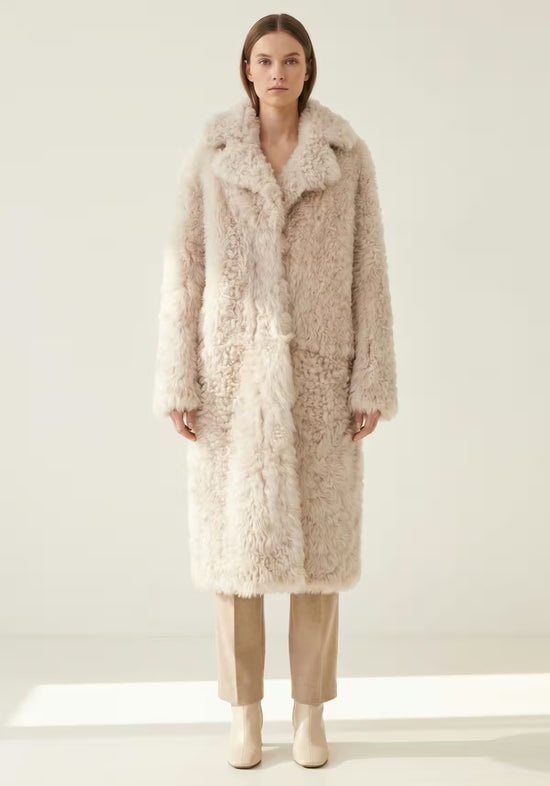 Women’s sheepskin coat made from curly sheepskin in cream-beige, full-length front view worn with trousers and boots.