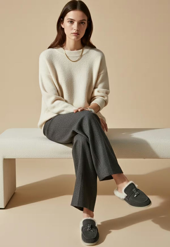 Women's sheepskin mules in anthracite shown in a seated styling shot with trousers and light wool socks.