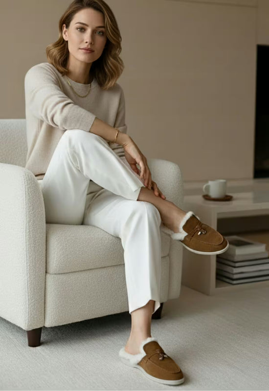 Brown ladies' sheepskin mules worn by a seated woman, with fur trim and buckle at the front.