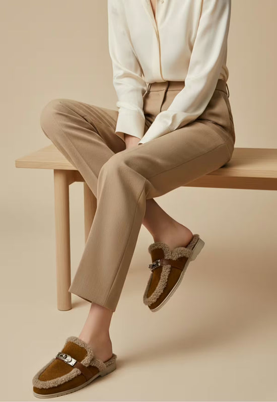 Brown women's sheepskin mules worn by a seated model with relaxed trousers.