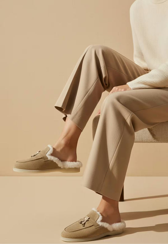 Women's sheepskin mules in sand worn side on, while seated against a plain background.