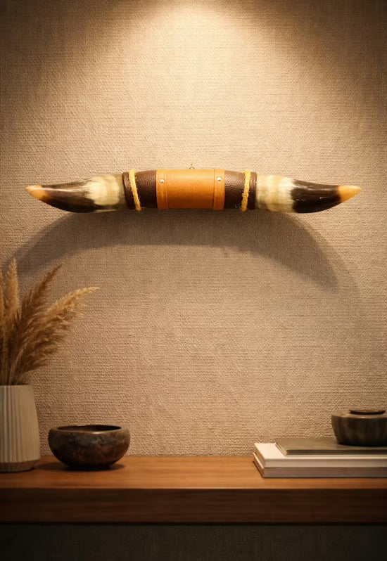 Wall decoration made of natural horn with leather and wood holder, front view staged above a shelf on a light wall.