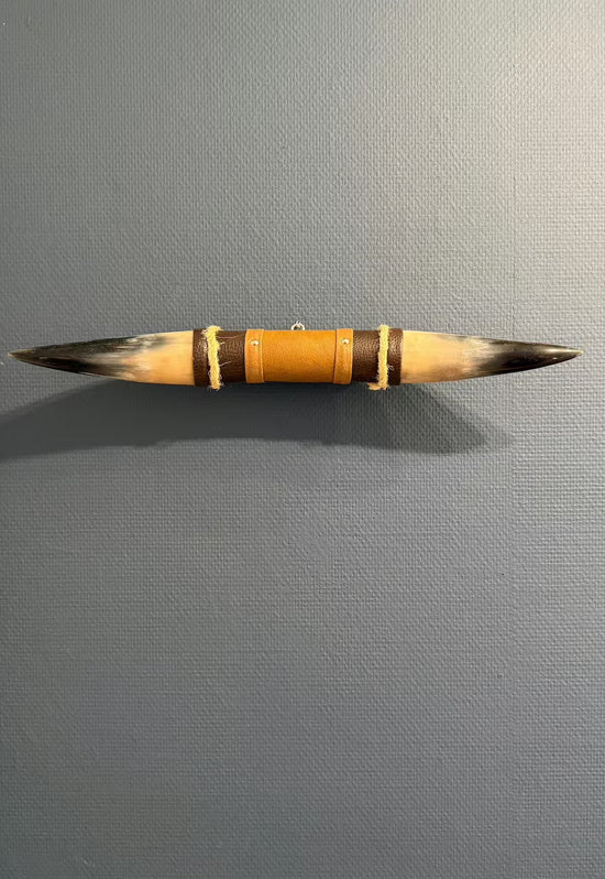 Wall decoration made of natural horn with leather and wooden bracket on a grey wall, shown frontally.