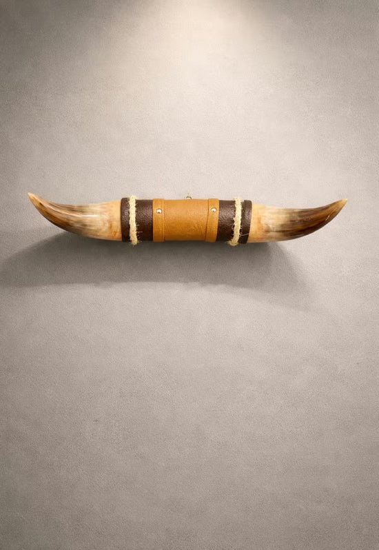 Horn wall object in brown with leather and wooden holder, shown front-on on a grey wall.